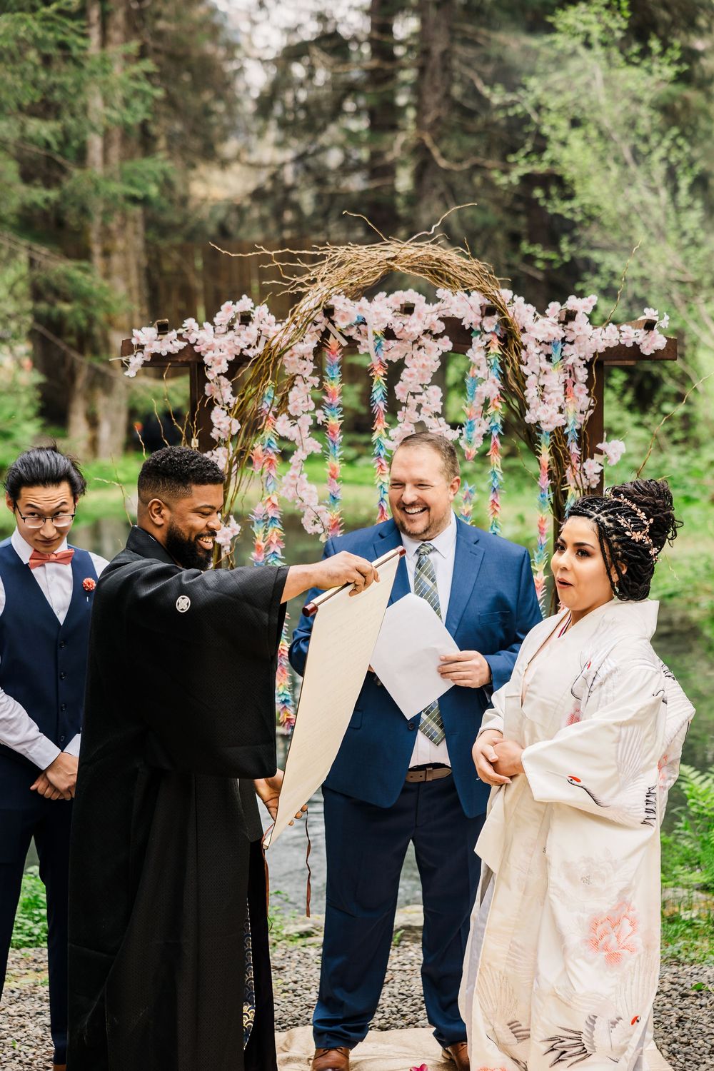 Wedding ceremony under a floral arch with pink blossoms in a wooded setting.