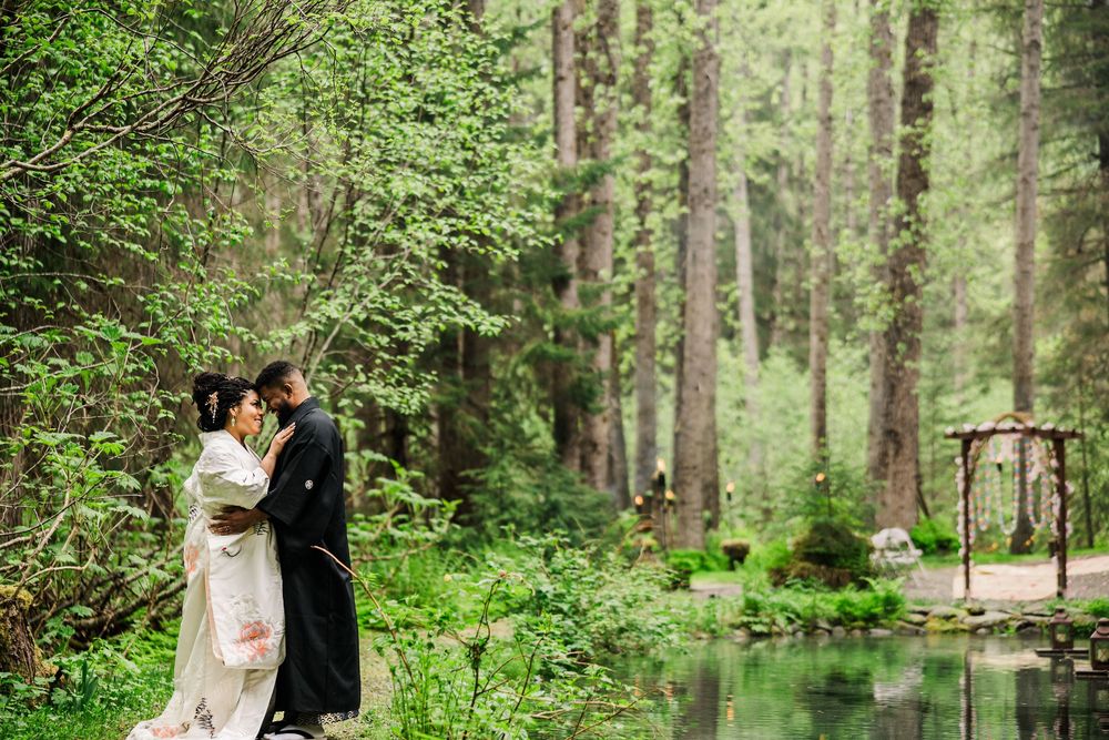 A romantic wedding portrait in a lush forest setting with a stream and wooden arch in the background.