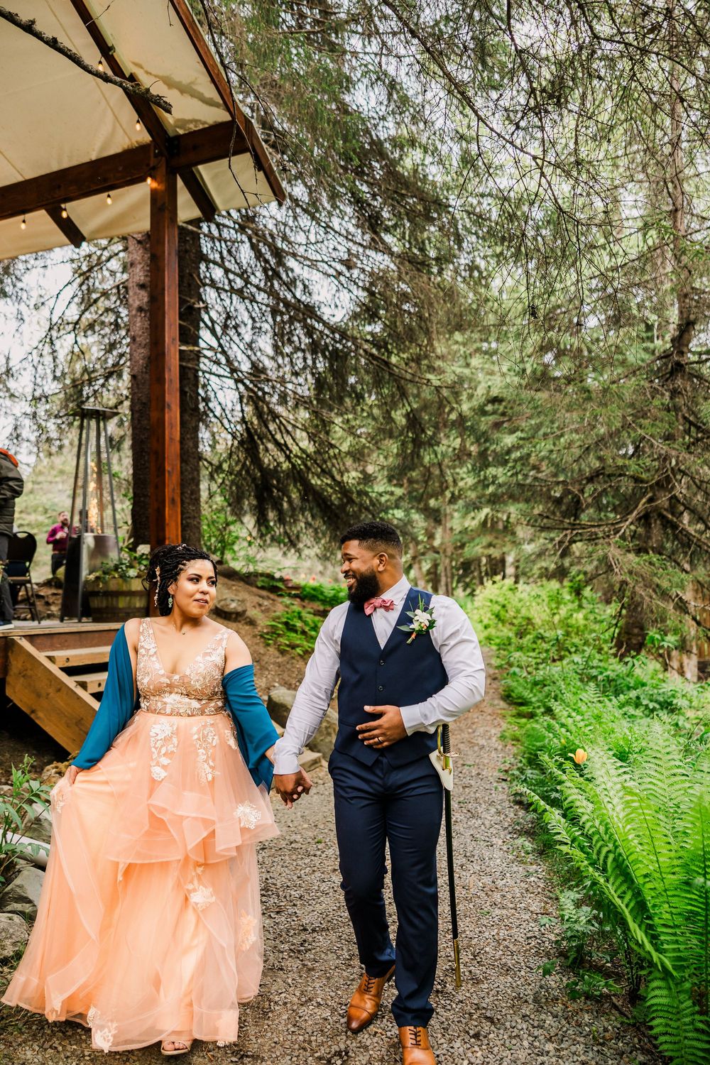 Couple walking hand in hand along a forest path near a rustic wooden structure.