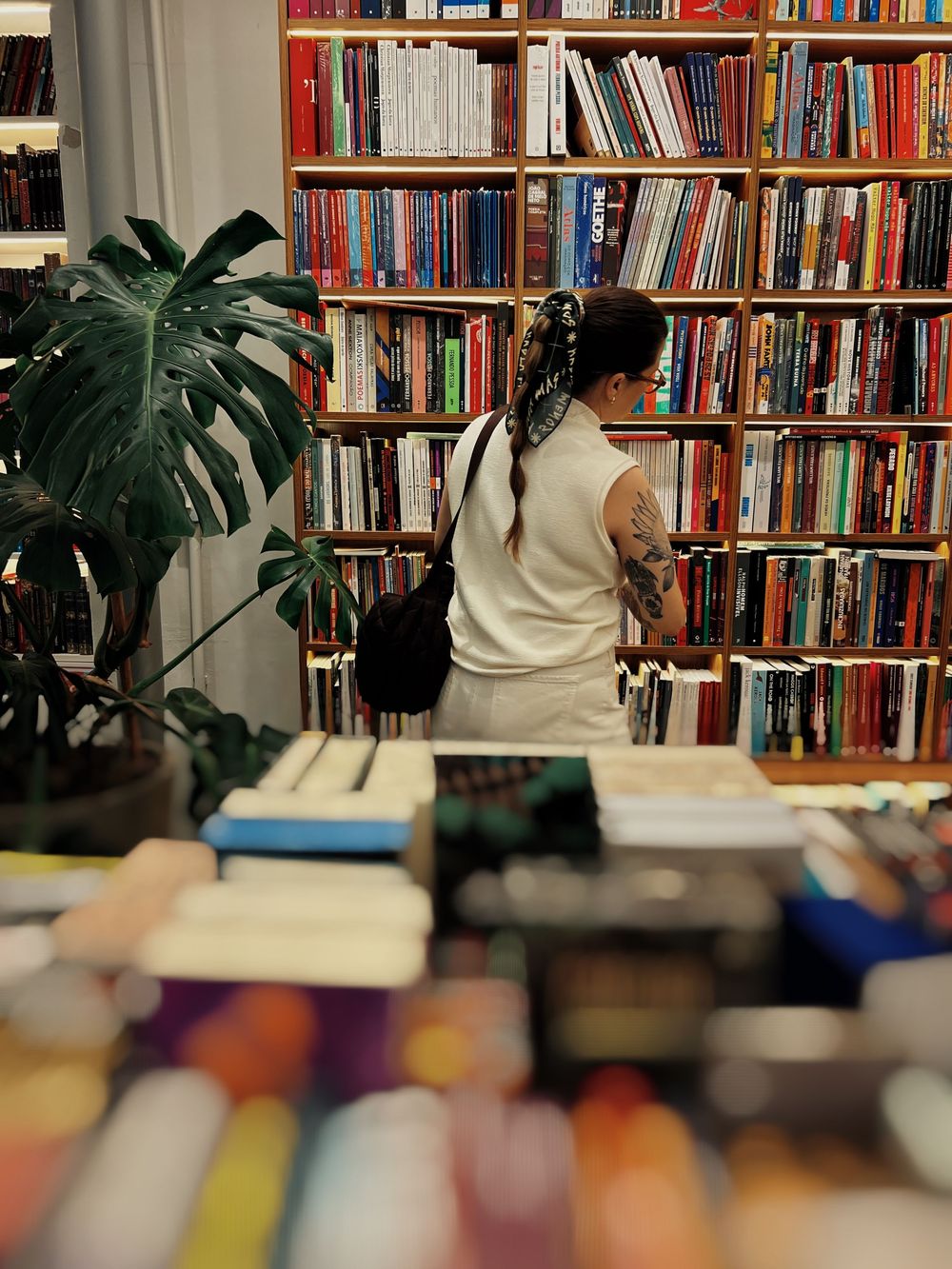 Person browsing in a cozy bookstore with floor-to-ceiling bookshelves and large indoor plants.