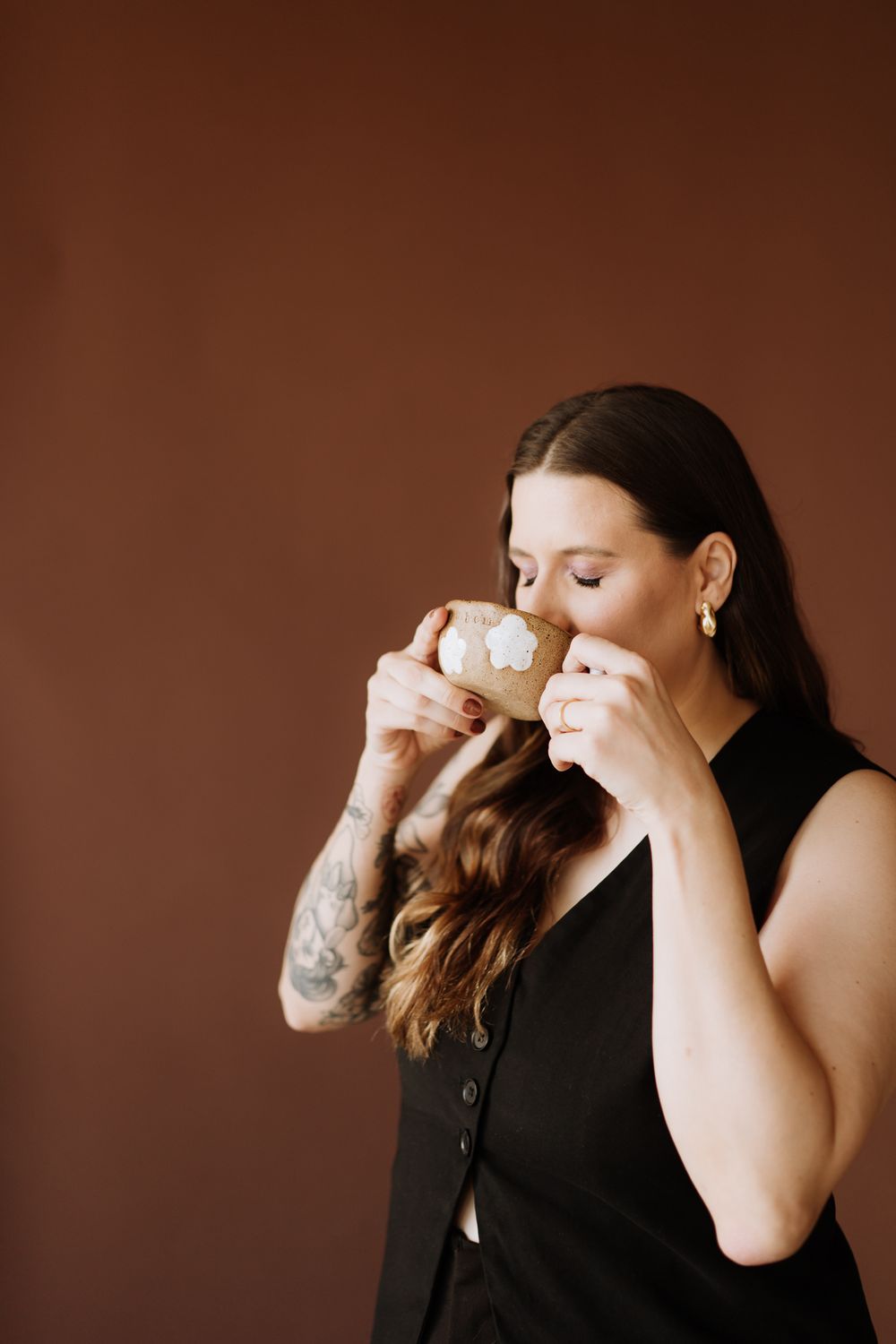 A figure in a black top takes a sip from a cup against a warm brown background.