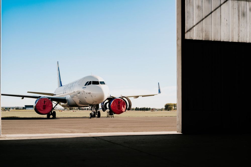 Commercial passenger aircraft with red engine cowlings sits at entrance of airplane hangar in bright daylight.