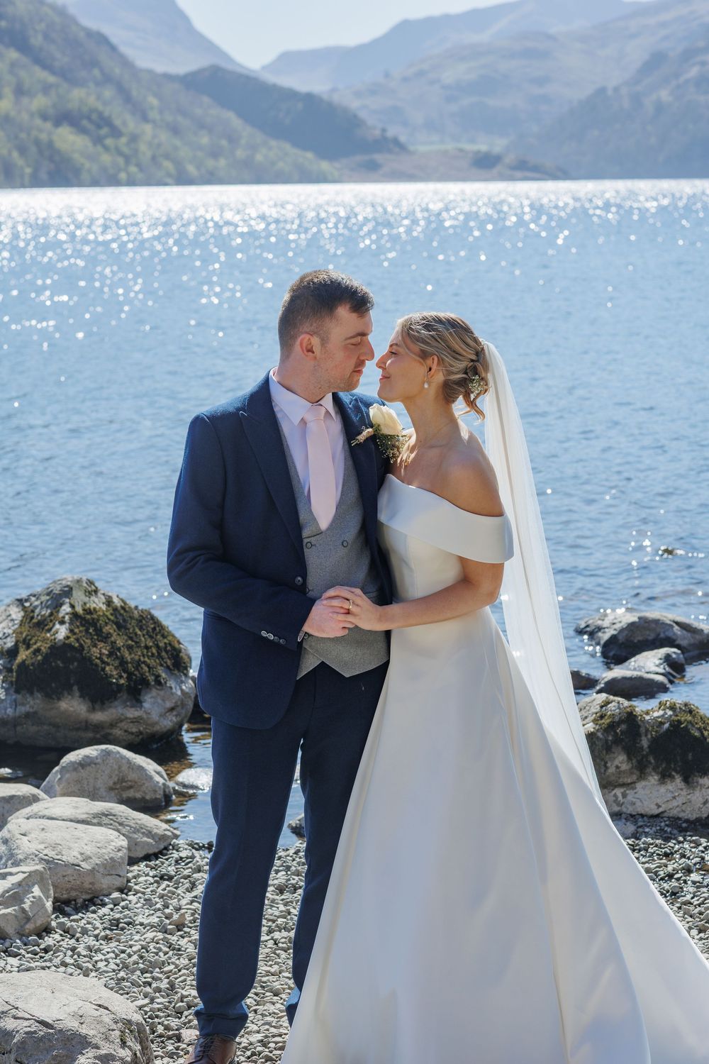 Wedding couple posing on rocky shoreline with mountains and lake in background.