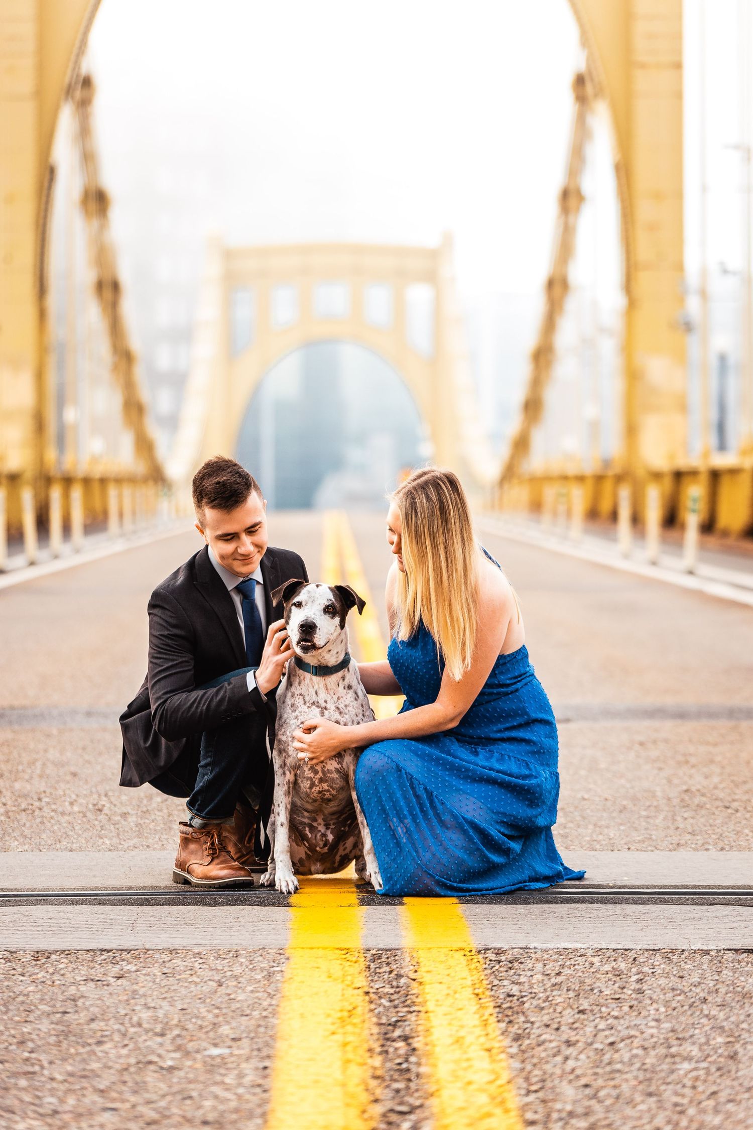 A couple kneels on a yellow-lined bridge with their spotted dog wearing a bowtie under warm golden lighting.