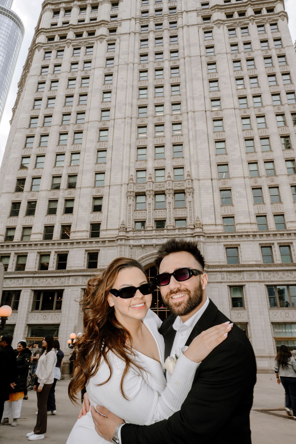 A couple wearing sunglasses embrace in front of a tall historic building in an urban setting.
