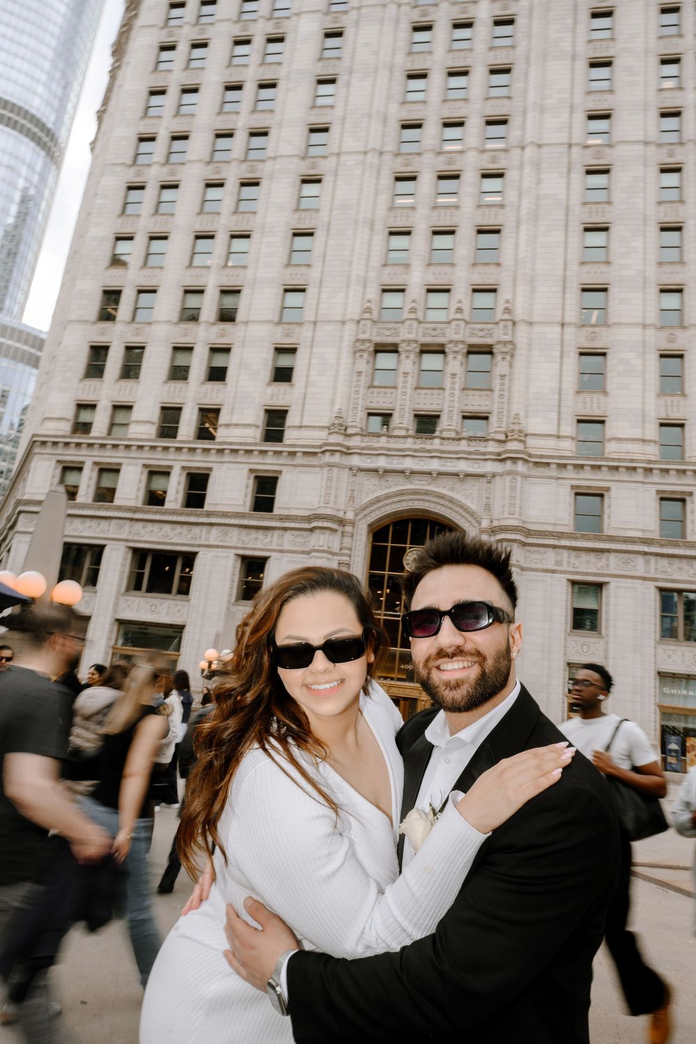 A stylish couple wearing sunglasses poses together outside a tall classical building on a sunny day.