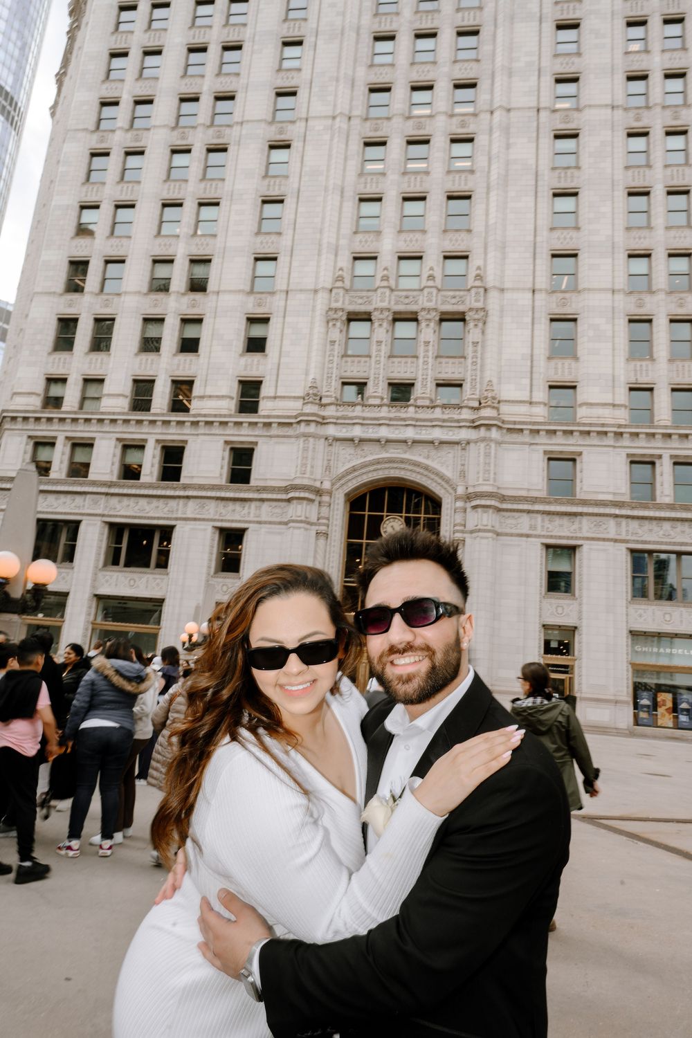 A couple wearing sunglasses poses together in front of a grand historic building in the city.
