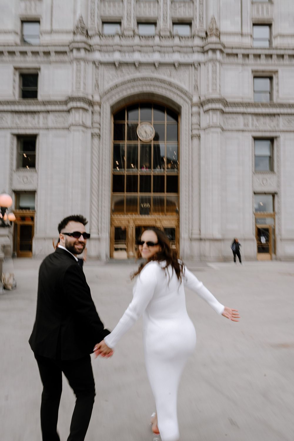 Newlyweds pose playfully in front of an elegant stone building entrance, with bride in white dress and groom in black suit.