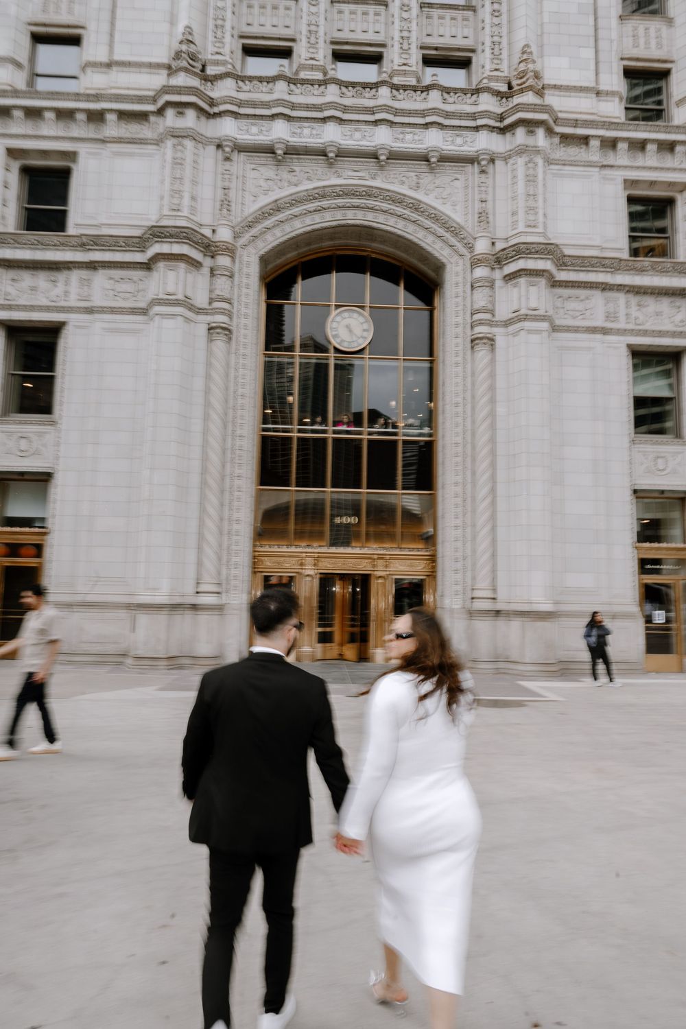 Couple walking towards ornate white stone building with Gothic architectural details and large arched entrance in the city.