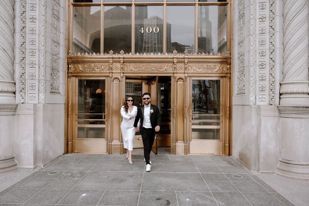 A couple exits through grand brass and glass doors of a historic building entrance.