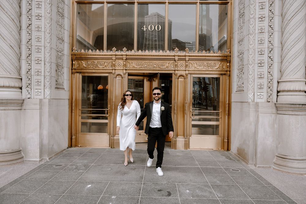 A couple walks out from an ornate building entrance with white stone columns and gold-trimmed glass doorway.