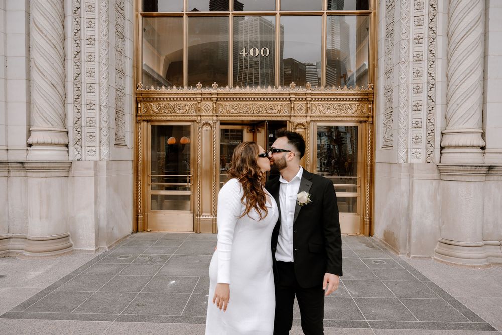 Two elegantly dressed people in wedding attire stand together in front of an ornate building entrance with classic columns.