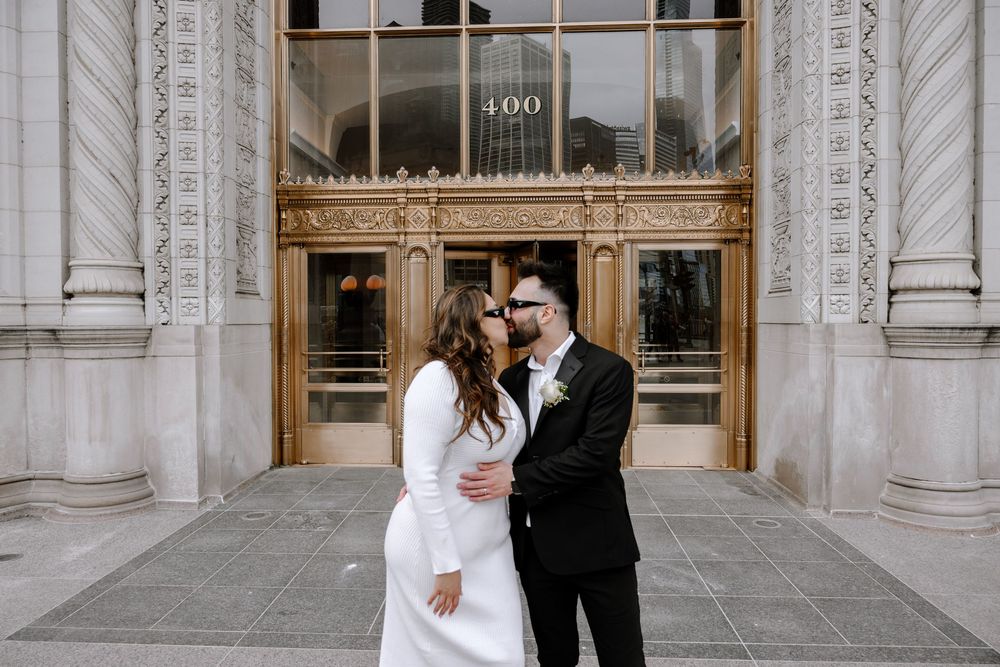 A couple shares a romantic moment in front of ornate stone columns and golden doors of a historic building.