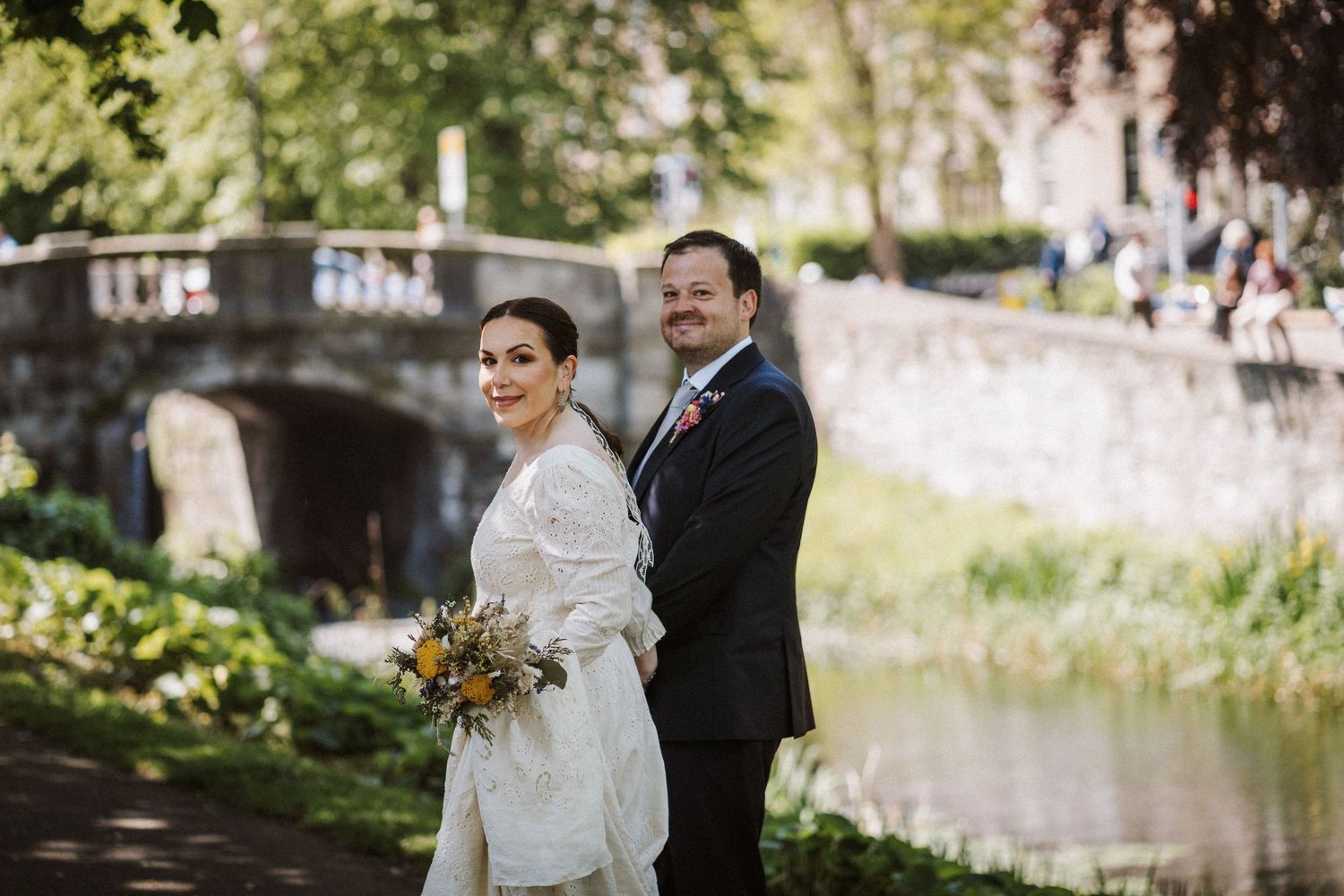 A couple stands together at a canal bridge in elegant wedding attire with autumn florals on a sunny day.