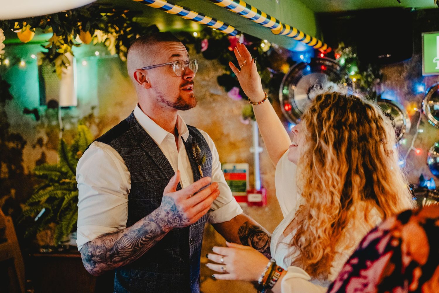 Cheerful couple dancing together at a colorful party venue with festive string lights and decorations in the background.