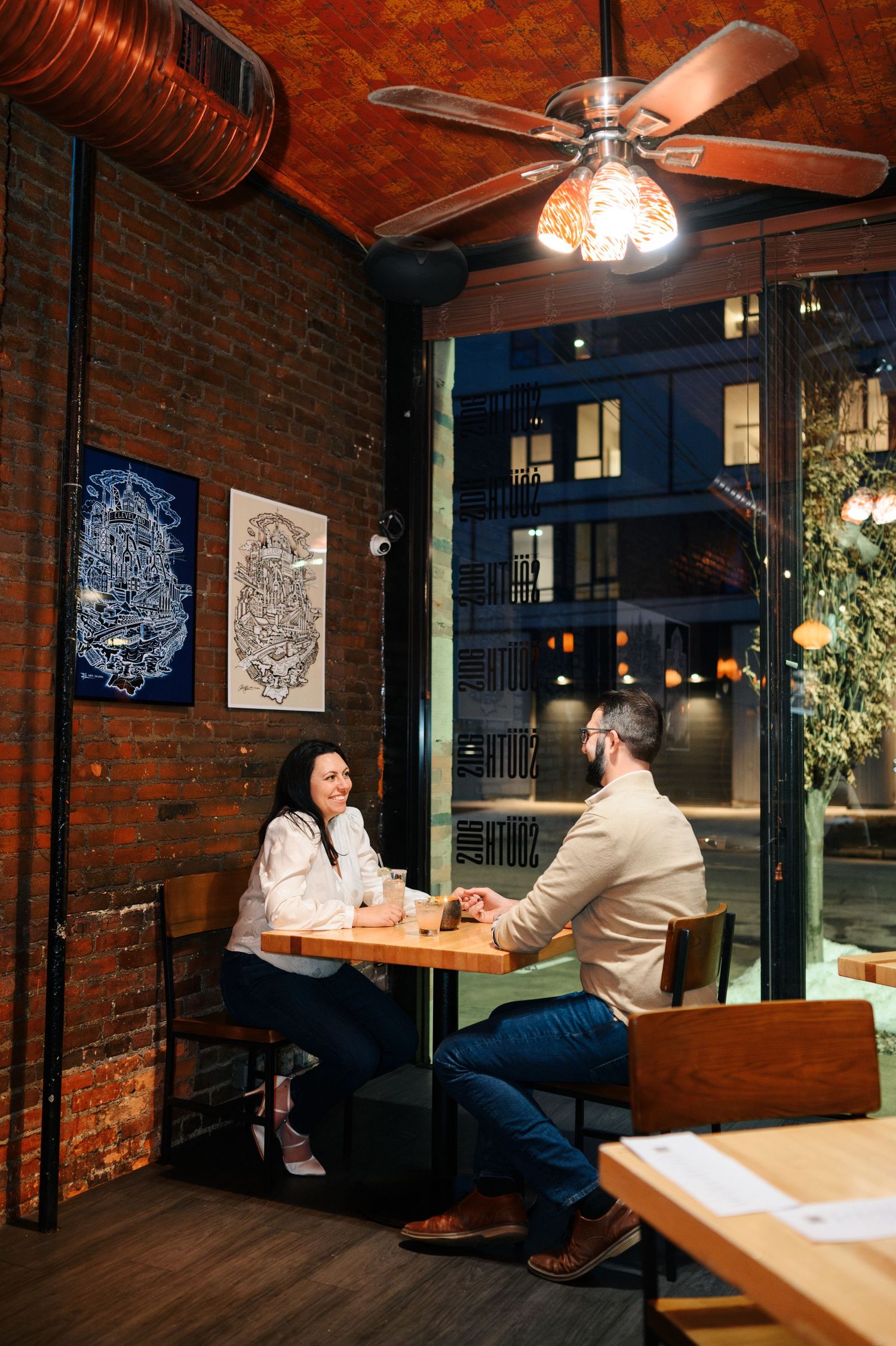 Two people enjoy drinks at a high-top table in a cozy restaurant with exposed brick walls and industrial ceiling details.