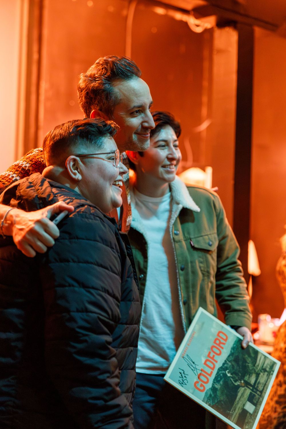 Three people standing together and laughing in warm orange lighting while one holds a book or document.