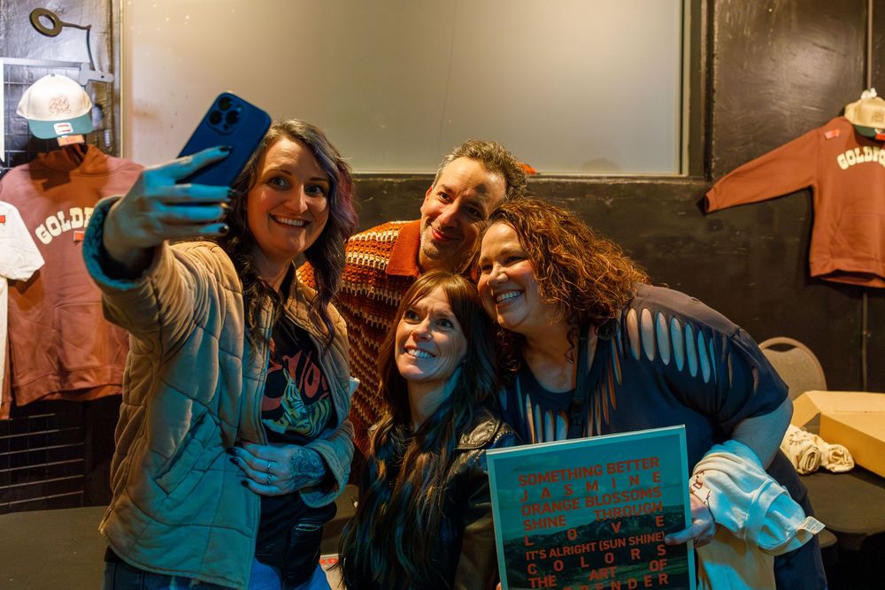 A group of friends takes a selfie together while smiling and posing with a book in a casual indoor setting.