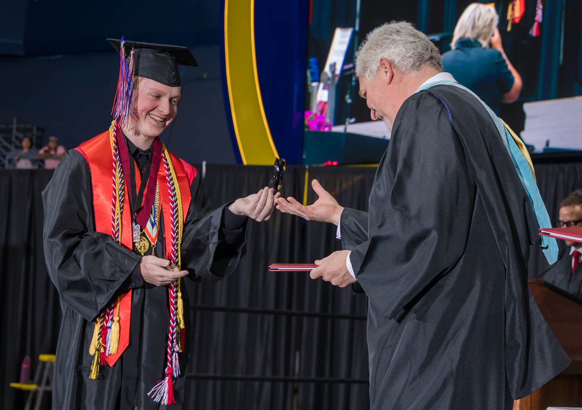 A graduate student in cap and gown receives their diploma from faculty member during commencement ceremony on stage.