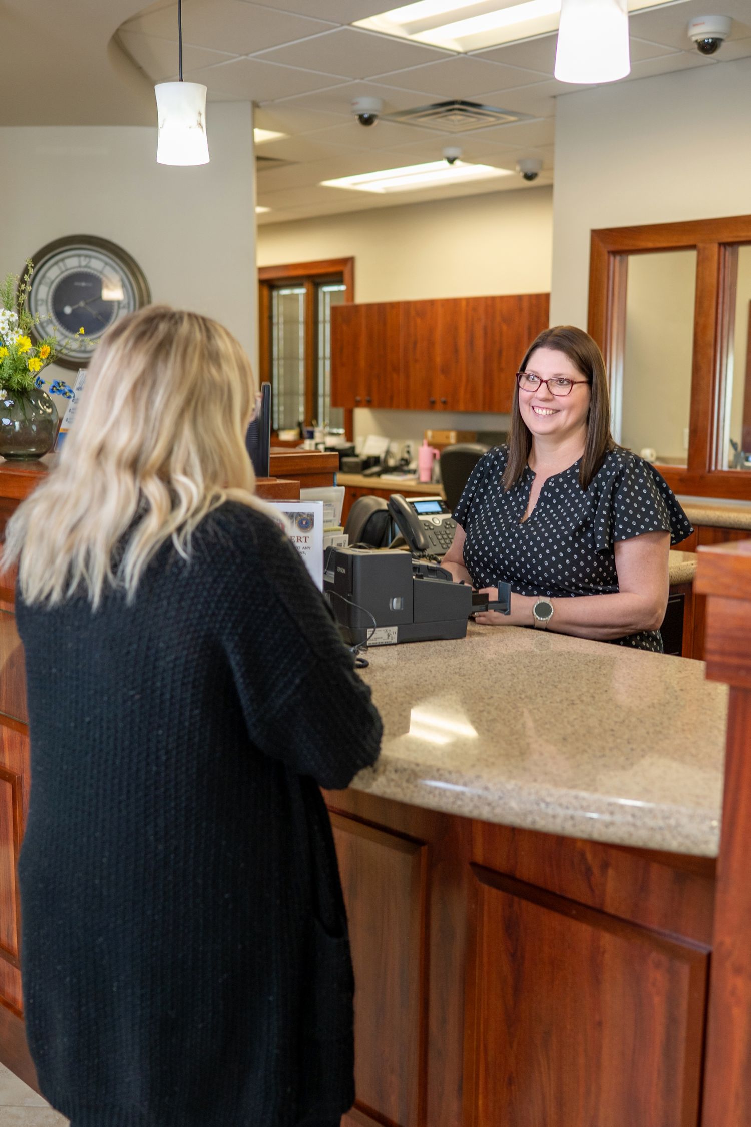 A bank employee assists a customer at a wood-paneled service counter beneath pendant lighting in an office interior.