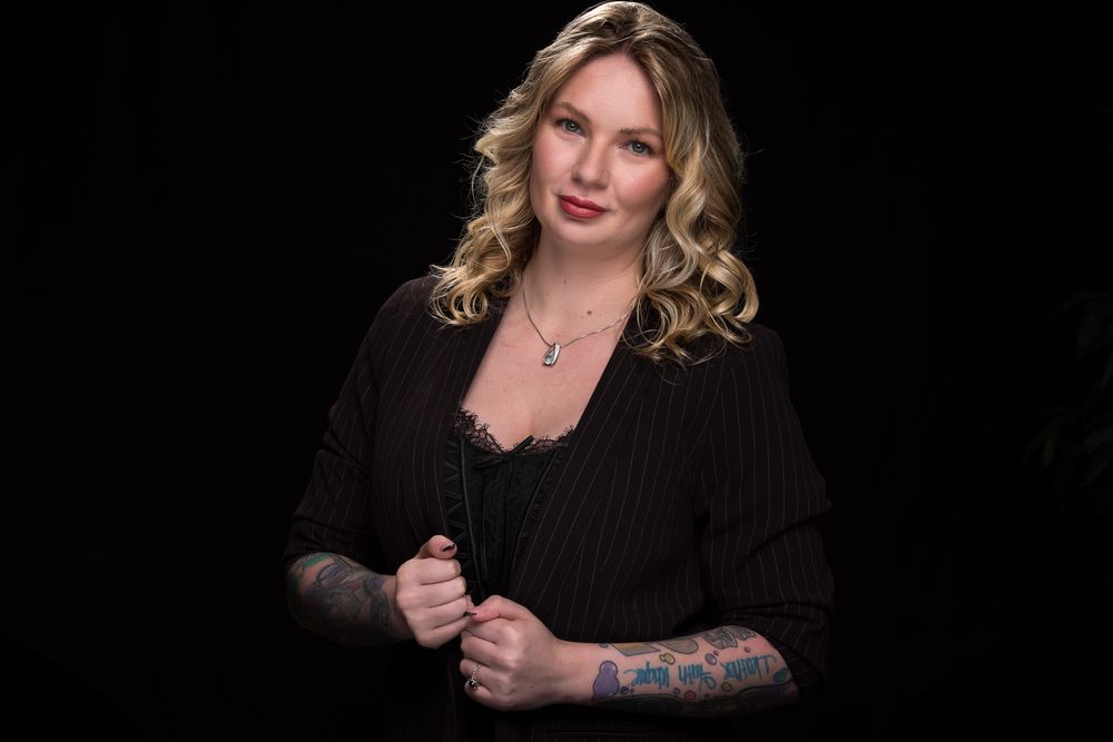 Studio portrait of a blonde woman wearing a black cardigan with visible arm tattoo against dark background.