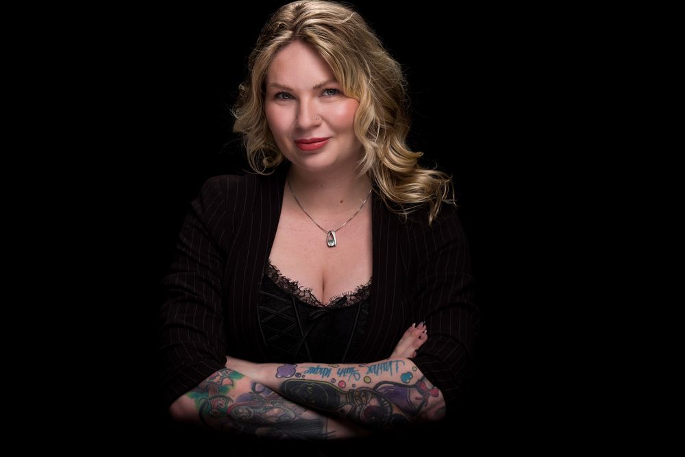 Studio portrait of a person wearing a black blazer with colorful tattoo sleeves against a dark background.
