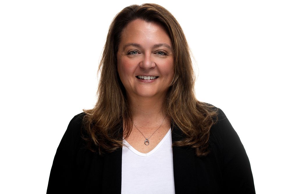 Professional headshot of someone in a white v-neck shirt and black blazer against white background with warm smile.