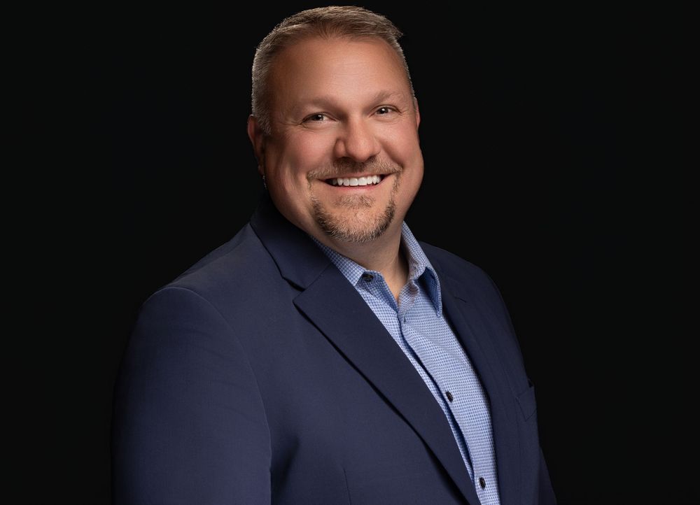 Professional headshot of someone in a navy blazer and light blue shirt against a dark background with a warm smile.
