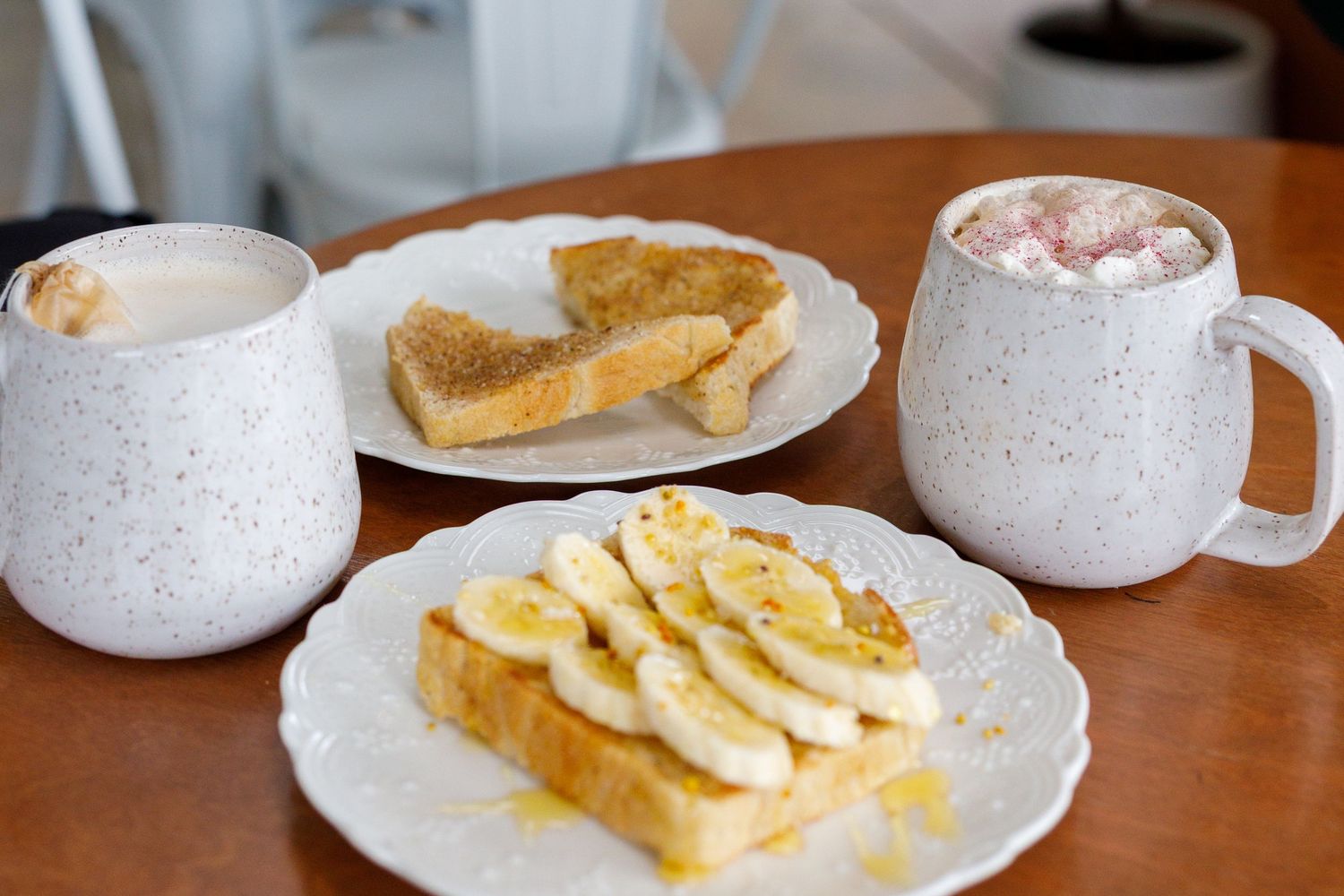 A cozy breakfast setup with two speckled white mugs and toast topped with sliced bananas and honey on white plates.