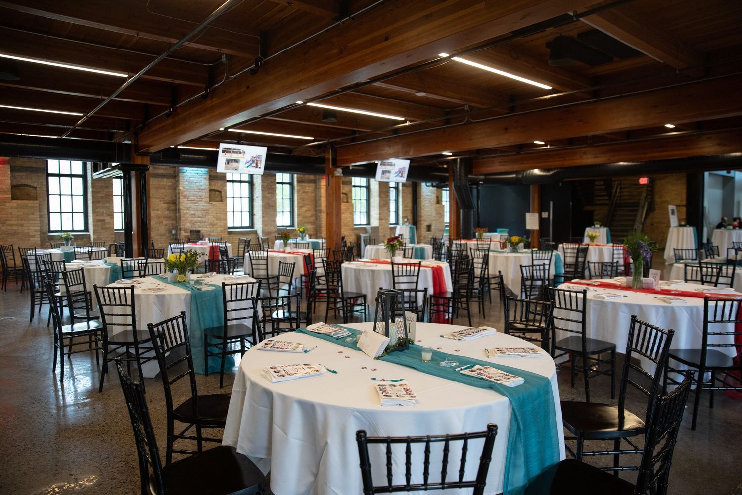 An elegant wedding reception venue with round tables set with white linens and dark Chiavari chairs under wooden beams.