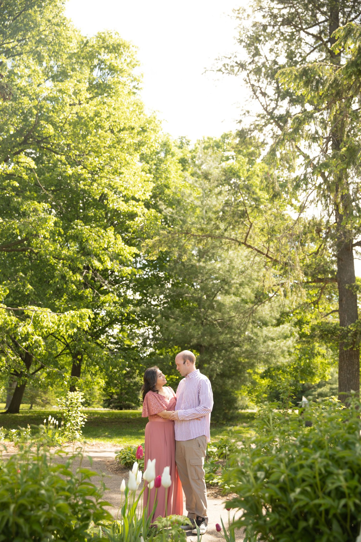 A couple shares a romantic moment on a sunlit garden path surrounded by lush green trees and white flowers.