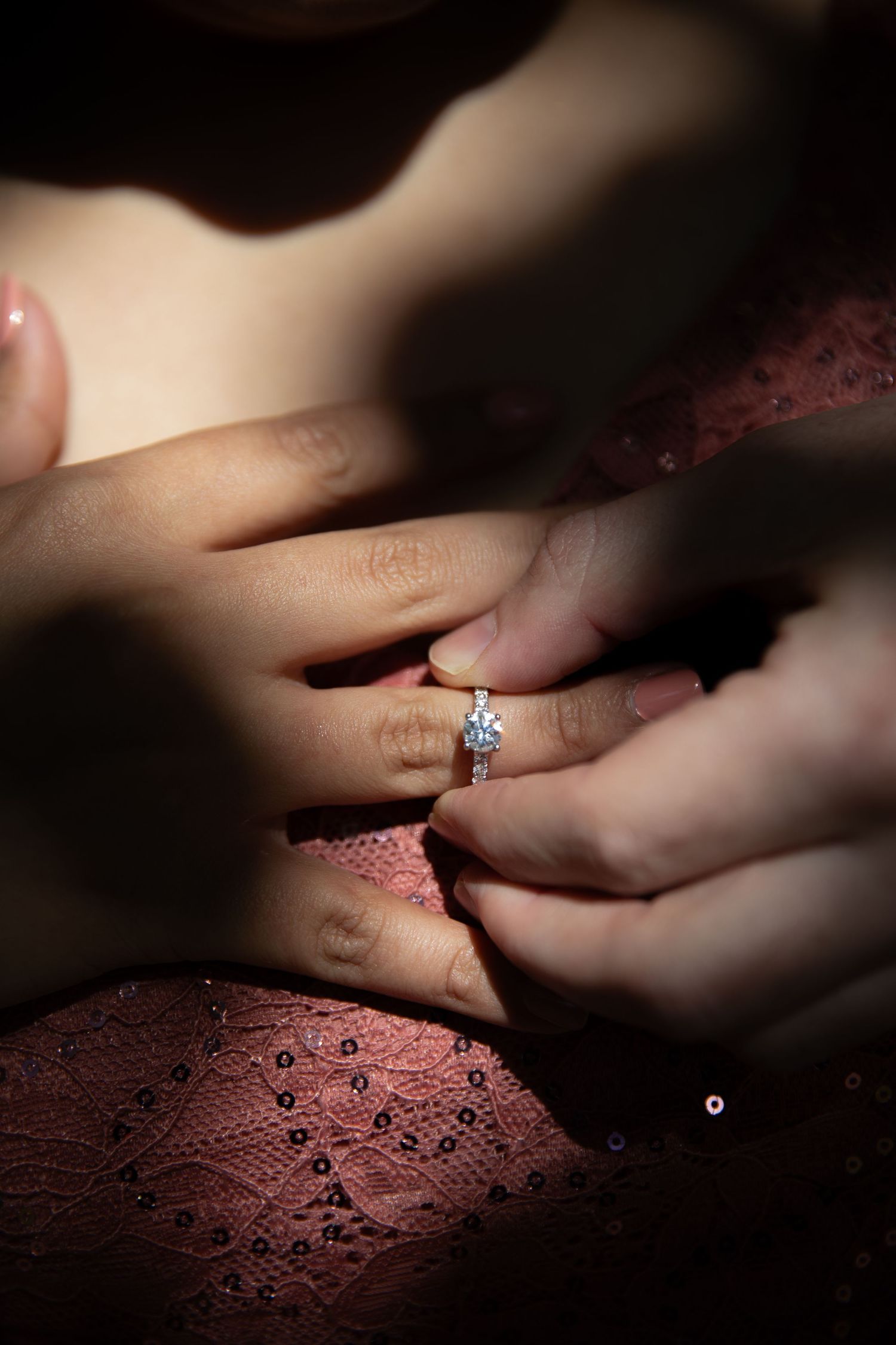 A close-up shot of hands placing an engagement ring with a sparkling diamond on a finger against pink lace fabric.