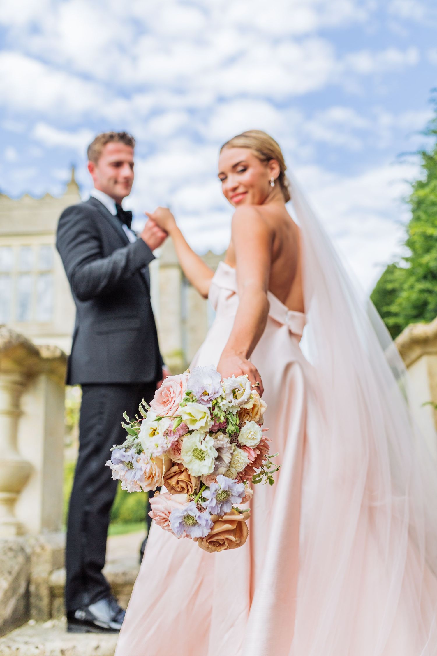 Bride in blush pink wedding gown holds elegant floral bouquet while standing on stone steps at Euridge - by Hayley Elizabeth Photography