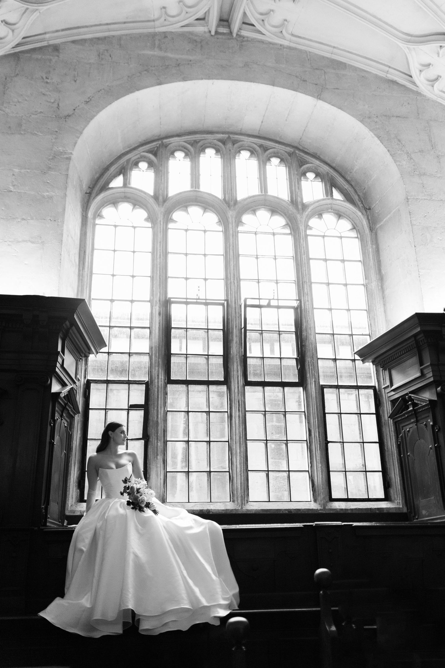 Black and white photograph of a bride in flowing white dress seated by window in dramatic silhouette at Bodelian Library Oxford - Hayley Eli