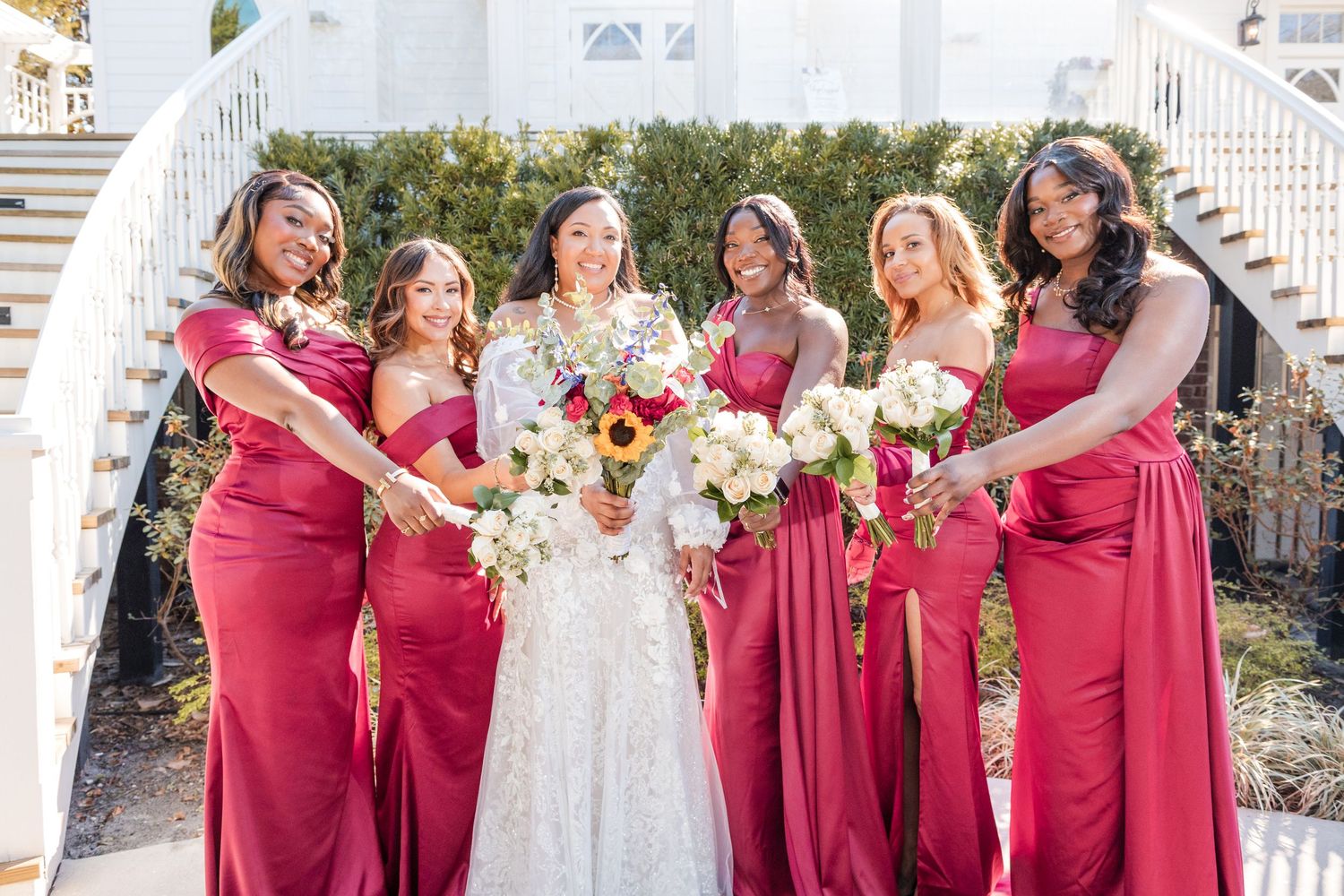 A wedding party wearing white and burgundy dresses poses together on outdoor steps with elegant floral bouquets.