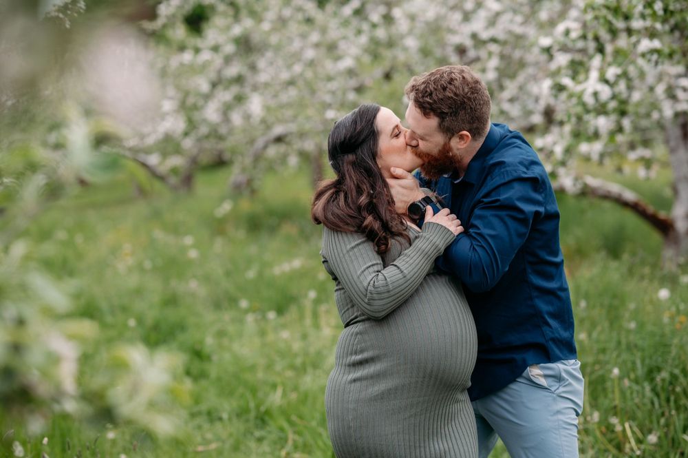 A couple shares a tender moment in a blooming spring orchard, with the woman wearing a grey maternity dress.