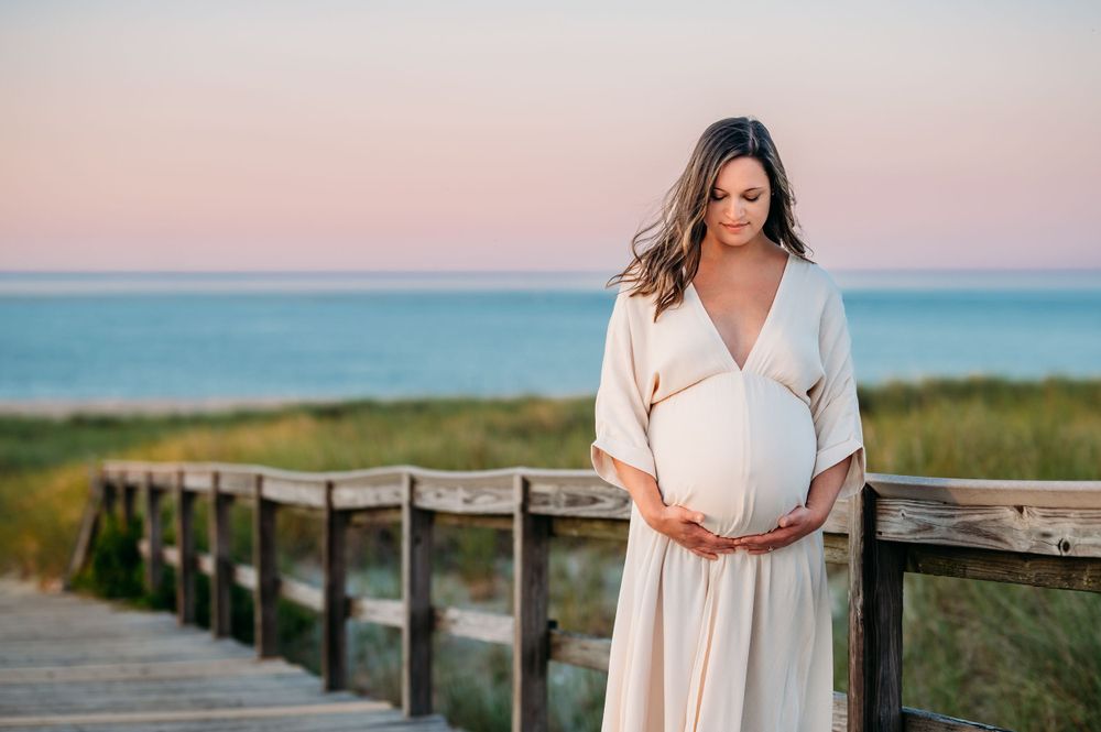 A woman in a flowing cream dress stands on a wooden boardwalk during sunset with ocean and sky visible behind her.