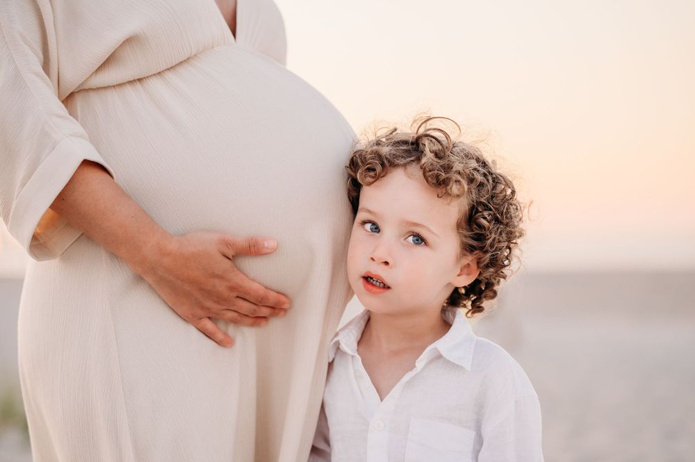 An adorable child with curly hair in white shirt gently touching a pregnant belly during an outdoor maternity photo session.