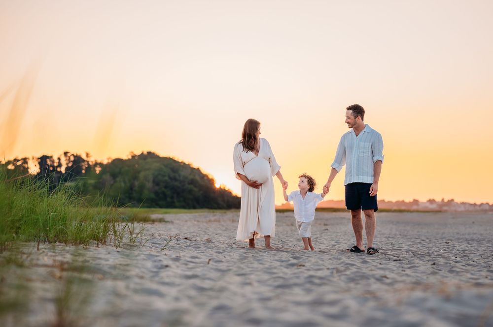 A family in white outfits walks together on a beach at sunset with tall grass in the background.