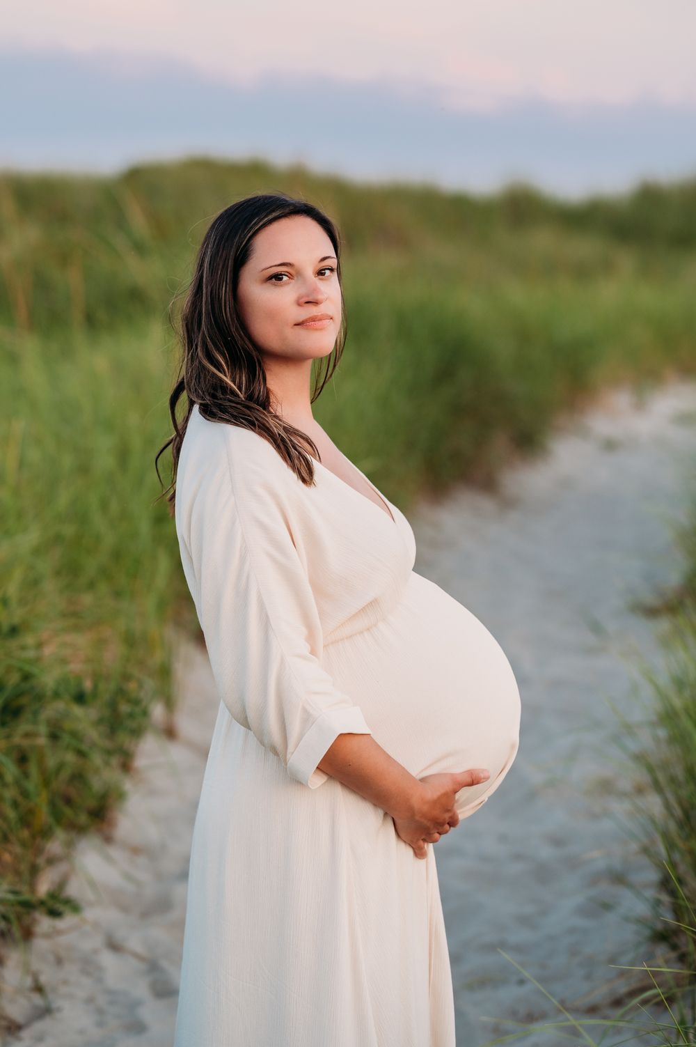 A woman in a flowing white dress stands on a sandy path at sunset cradling her pregnant belly.