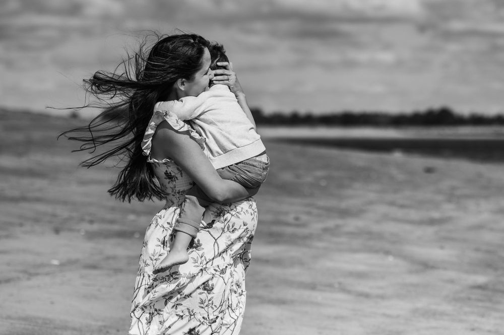 A person with long flowing hair embraces another in a floral dress on a windy beach in black and white.