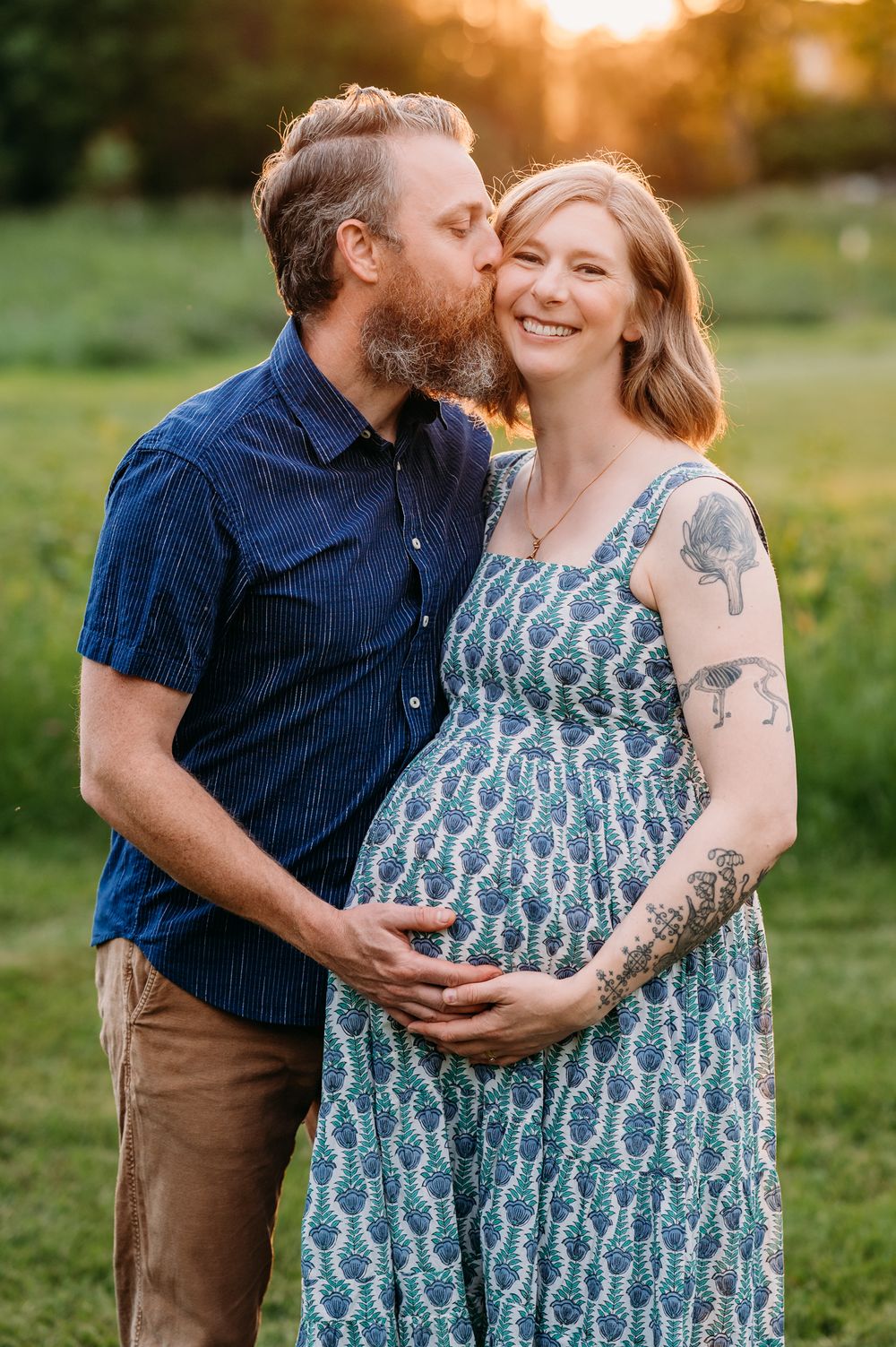 A loving couple shares a tender moment at sunset in a grassy field, with her wearing a patterned blue maternity dress.