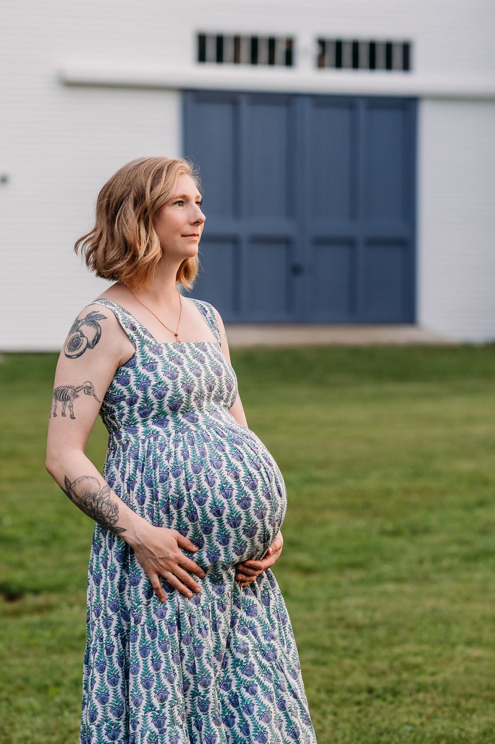 Expectant parent in patterned blue sundress poses outdoors on grass with hand on belly.