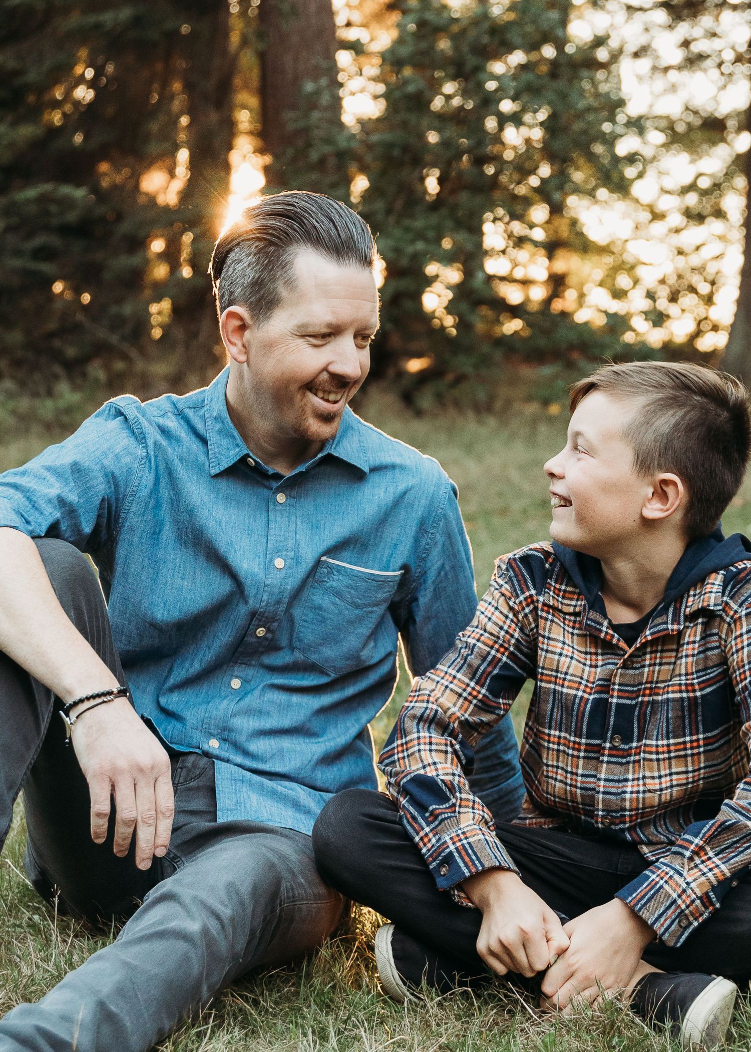A tender moment captured between two people sitting on grass during golden hour in a forest setting.