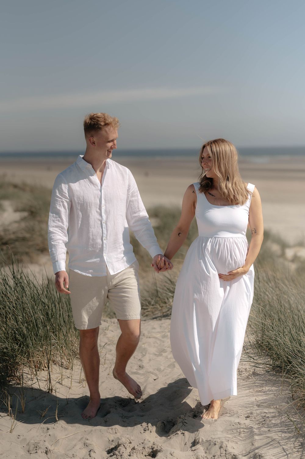 A couple in white outfits holding hands while walking on sand dunes at the beach during a maternity photoshoot.