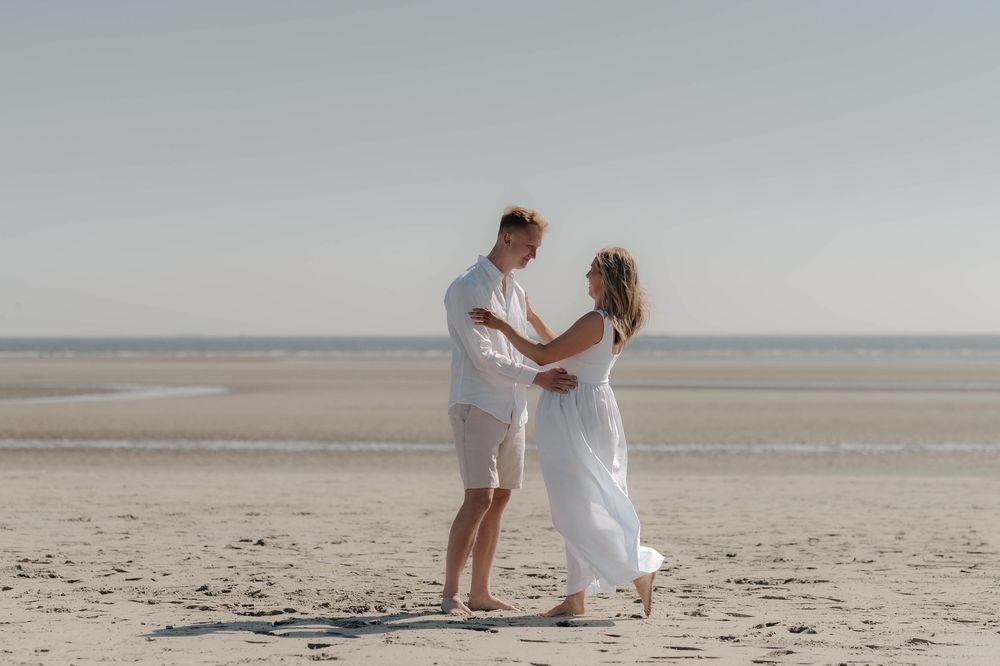 A couple dances together on a serene sandy beach with calm waters and clear skies in the background.