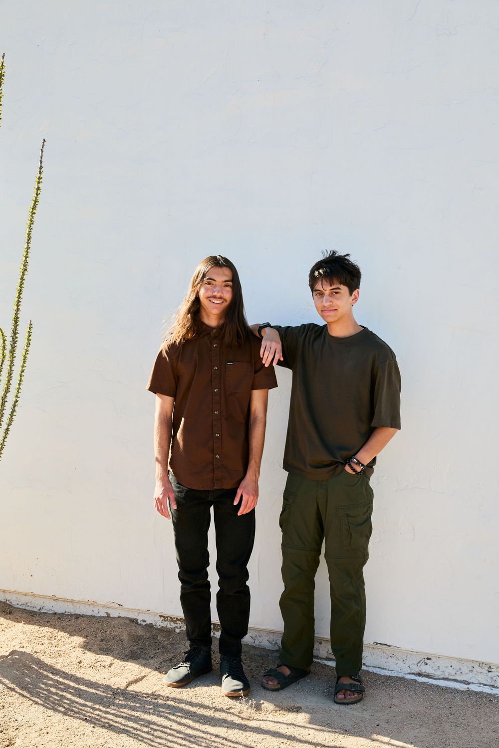 Two people in casual attire stand against a white wall with a tall cactus plant visible in the corner.