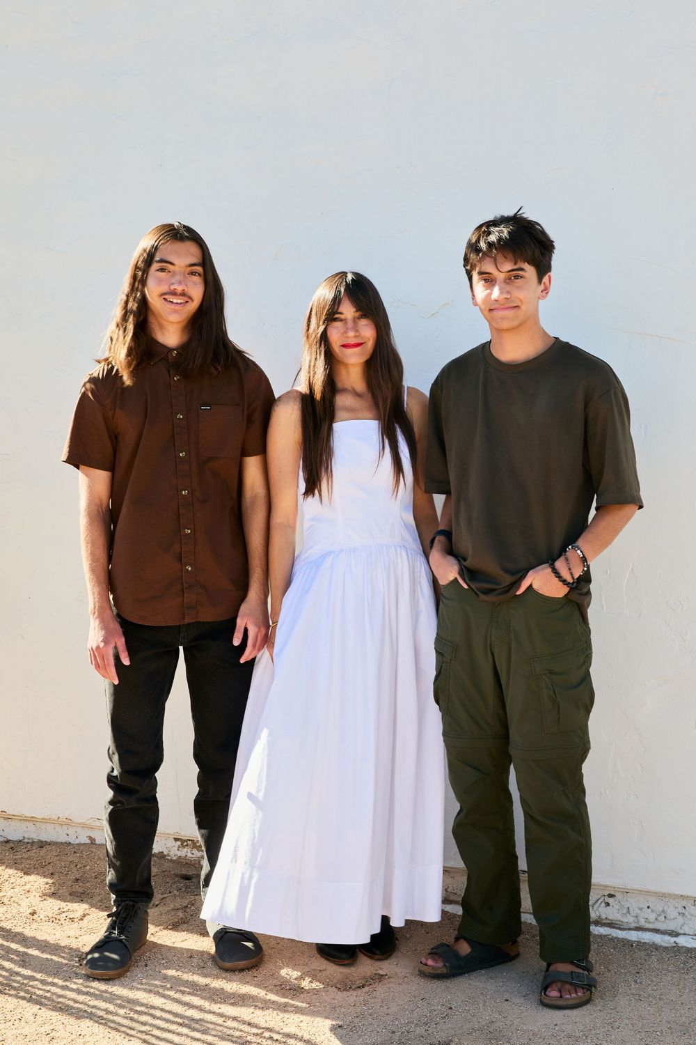Three friends stand together wearing contrasting outfits of brown, white and black in a casual outdoor setting.
