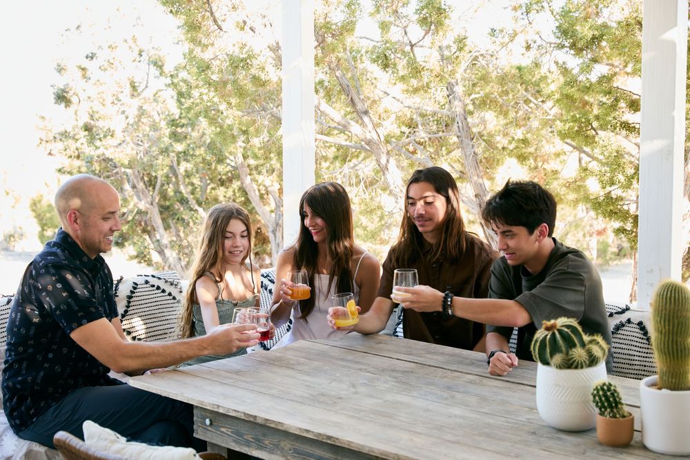 A group of people share drinks and conversation while gathered around a rustic wooden table on an outdoor porch.