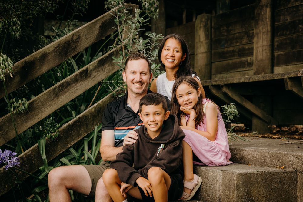 A family of four sits together on wooden steps outdoors, smiling at the camera in casual summer attire.